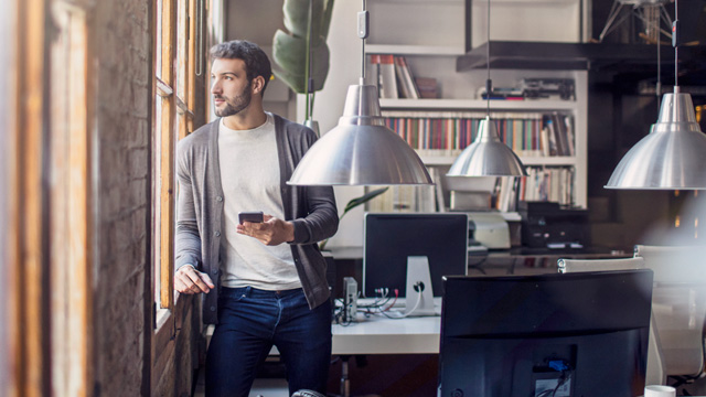 Man in an office thinking when looking outside and holding a mobile